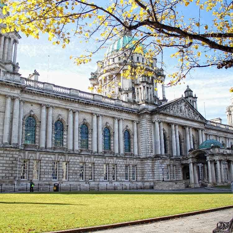 Belfast City Hall under a clear blue sky, viewed from the lawns with golden autumn leaves framing the historic architecture.