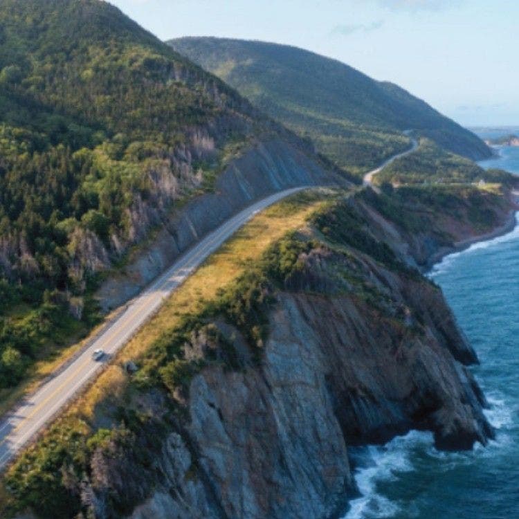 An aerial view of the Cabot Trail winding along the rugged, forested cliffs of Cape Breton Island, Canada, for the Transatlantic Crossing section.
