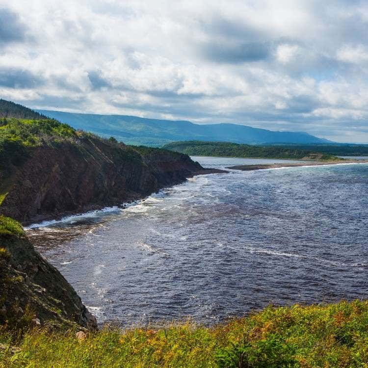 Waves crashing against the rugged rocky cliffs of a scenic green coastline where a river channel meets the open ocean.