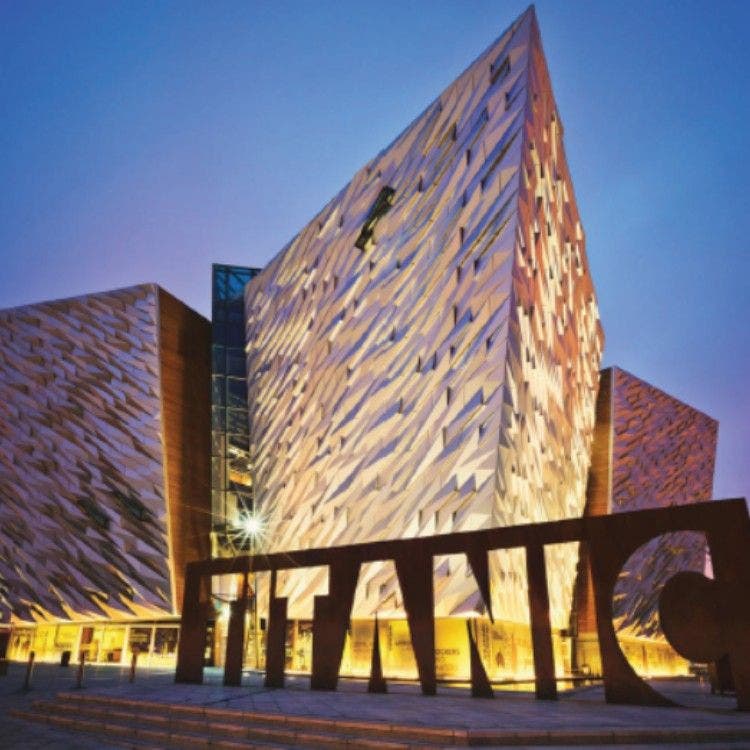The striking, illuminated geometric facade of the Titanic Belfast museum at dusk, with the large rusty steel "TITANIC" sign in front.