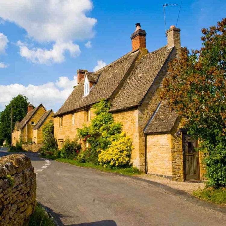 Charming honey-colored stone cottages with sloped roofs lining a quiet village street in the English Cotswolds.