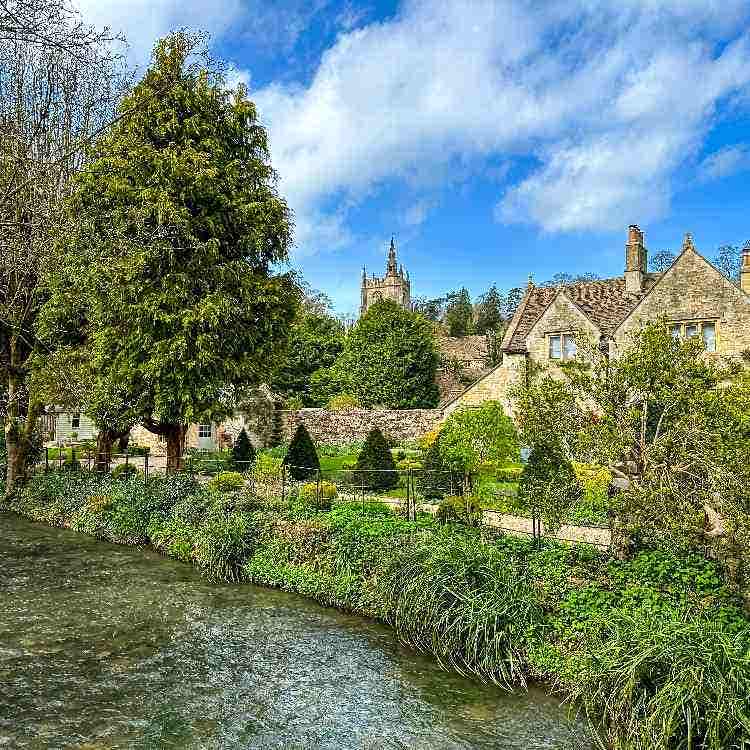An English stone cottage and lush garden beside a flowing stream in the Cotswolds, with a church tower behind.