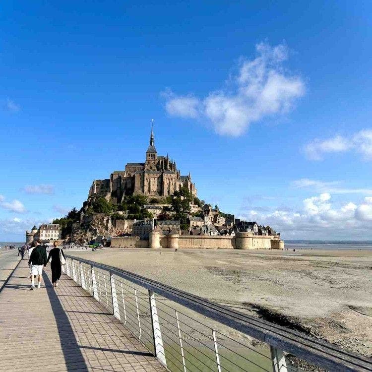 Visitors walking along a modern footbridge toward the medieval tidal island of Mont-Saint-Michel under a bright blue sky.