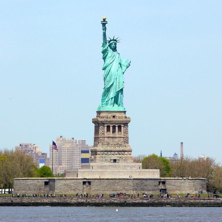 The iconic Statue of Liberty standing tall on its large stone pedestal on Liberty Island, with buildings visible in the background.